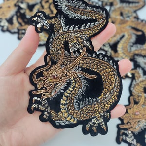 May include: A gold and silver embroidered dragon applique patch with sequins. The dragon is curled up with its head facing forward and its wings spread out.