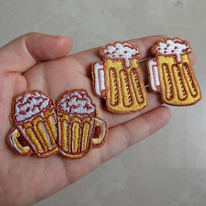 May include: Embroidered patches depicting beer mugs with white frothy foam. The mugs are yellow and brown with white detailing. These patches are designed for application on clothing or accessories, offering a playful aesthetic.