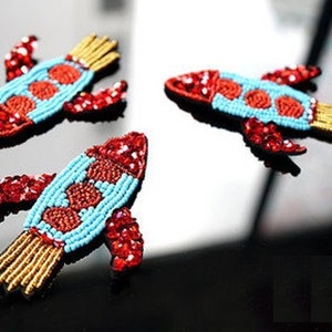 Könnte beinhalten: Drei gestickte Raketen-Patches mit roten, blauen und goldenen Details. Die Raketen sind aus Perlen und Pailletten gefertigt.