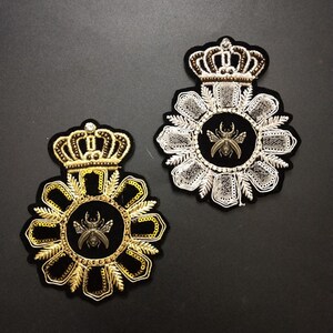Puede incluir: Dos parches bordados con una corona y un diseño de escarabajo. Un parche es dorado y negro, el otro es plateado y blanco.