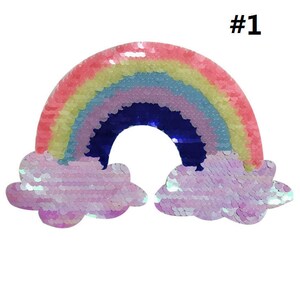May include: A rainbow-colored sequin patch with clouds on either side. The rainbow is made of pink, yellow, light blue, purple, and dark blue sequins. The clouds are white with iridescent sequins. The patch is labeled #1.