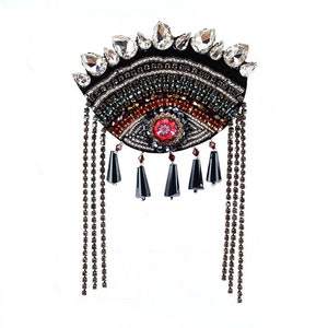 May include: A black eye-shaped brooch with colourful beaded details and a red centre. The brooch has a fringe of silver chains.