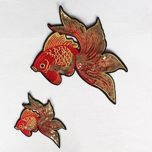 May include: Two embroidered goldfish patches. The fish are red with gold and sequin accents. The patches have black borders and are set against a white background. One patch is larger than the other.