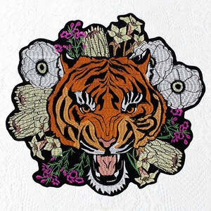 May include: Iron-on patch embroidered with a tiger's face, featuring an open mouth and surrounded by white, yellow and pink flowers. The tiger has orange and black stripes.