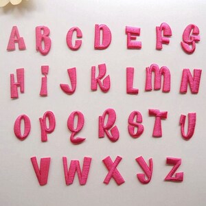 Op de afbeelding: Een set van 26 roze viltletters, van A tot Z, in een cursief lettertype. De letters zijn individueel uitgesneden en klaar om te worden gestikt of gestreken op stof.