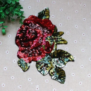 Könnte beinhalten: Eine große, rote, paillettenbesetzte Rose mit grünen, paillettenbesetzten Blättern. Die Rose ist ein dekorativer Schmuck für Kleidung oder Bastelarbeiten.