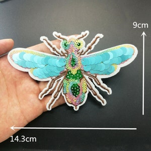 Può includere: Un'applicazione di ape colorata con paillettes blu iridescenti sulle ali. L'ape è alta 9 cm e larga 14,3 cm.