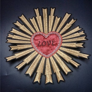 May include: Embroidered patch featuring a red velvet heart with the word "LOVE" in black script. The heart is surrounded by a beaded border and gold rays on a black background. This decorative patch is suitable for clothing or accessories.