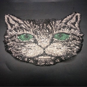 Op de afbeelding: Een zwarte en zilveren pailletten kattengezicht applique met groene ogen.