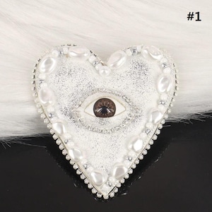 May include: Heart-shaped brooch featuring a central eye design. The brooch is white with a glittery texture, framed by faux pearls and small silver beads. The eye is brown with a white background. The number "#1" is in the upper right corner.