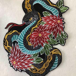 May include: Embroidered patch featuring a black snake with teal and gold accents, intertwined with red and yellow flowers and green leaves. The patch has a detailed design and is likely for clothing or accessories.
