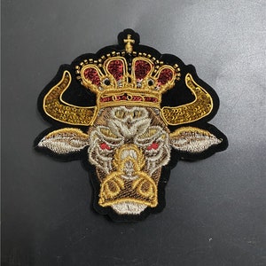 May include: Embroidered patch featuring a bull's head wearing a gold and red crown. The bull has gold horns and red eyes, set against a black background. The patch is likely for clothing or accessories.