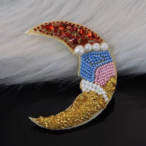 May include: A crescent moon brooch with a beaded design. The top section features red gems and gold trim. The middle section has blue, pink, and white beads. The bottom section is covered in gold beads. The brooch is set against a white, textured background.