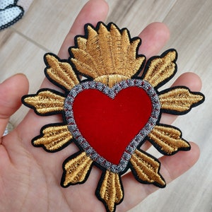 May include: A gold embroidered heart with a red velvet center and a gold halo. The heart is surrounded by a gold halo with a silver trim.