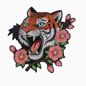 May include: Embroidered patch featuring a roaring tiger with orange and white fur, teal eyes, and a pink tongue. The tiger is surrounded by pink flowers with yellow centers and green leaves, set against a black background.