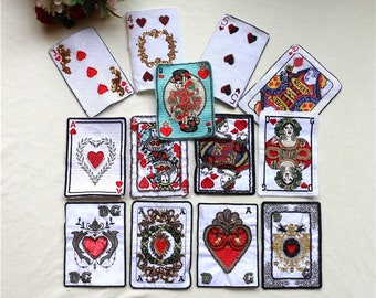May include: Twelve embroidered iron-on patches featuring playing cards with hearts, queens, and aces. The patches are white with red, gold, and black accents. The patches are designed in a vintage style.
