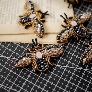 May include: Four beaded bee brooches with black and gold accents and pink centres. The brooches are arranged on a black and white grid.