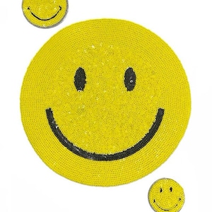Puede incluir: Tres parches amarillos con forma de cara sonriente con contornos negros hechos de lentejuelas. El parche más grande tiene un diámetro de unos 15 cm.