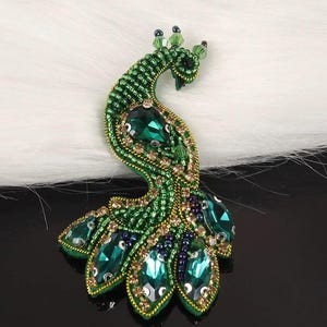 May include: A detailed peacock brooch, predominantly green with emerald-green crystal embellishments. The brooch is crafted with small beads and gold accents, creating a decorative and eye-catching accessory.