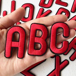 以下が含まれることがあります： 黒い縁取りのある赤い刺繍生地の文字A、B、C。白い背景に表示され、他の赤い文字も見えます。これらの文字は、おそらく衣類や工芸品用のパッチです。