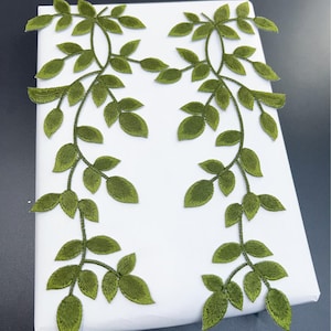 Pode incluir: Duas vinhas de folhas verdes bordadas com folhas detalhadas. As vinhas são dispostas simetricamente sobre uma superfície branca, criando um design decorativo. As folhas têm uma aparência texturizada, adicionando profundidade.