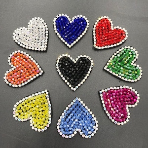 Könnte beinhalten: Eine Sammlung von neun herzförmigen Verzierungen. Jedes Herz hat eine andere Farbe, darunter Weiß, Blau, Rot, Orange, Schwarz, Grün, Gelb und Rosa. Die Herzen sind mit kleinen, funkelnden Kristallen bedeckt und mit klaren Strasssteinen umrandet.