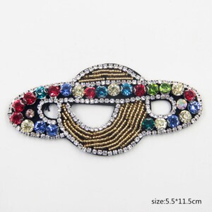 May include: A gold, black, and multi-colored beaded and rhinestone applique in the shape of a planet. The applique is 5.5 inches by 11.5 inches.