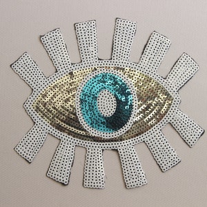 May include: A gold and turquoise eye with white sequins around it. The eye is a popular symbol of protection and good luck.
