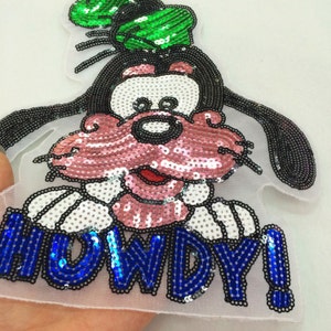May include: A sequined patch featuring a cartoon dog with a pink mouth and nose, black ears, and a green hat. The word "HOWDY!" is spelled out in blue sequins.