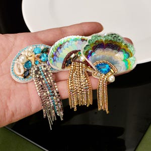 May include: Three decorative brooches featuring fan-shaped designs adorned with iridescent sequins, colorful crystals, pearls, and small seashells. Each brooch includes long, dangling fringe strands made of sparkling rhinestones in various metallic tones.