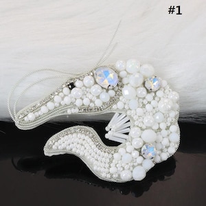 Puede incluir: Broche artesanal con forma de caballito de mar, adornado con cuentas blancas y cristales iridiscentes. El diseño incluye cuentas de diferentes tamaños y detalles plateados. El número "#1" es visible.