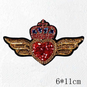 Pode incluir: Um patch em forma de coração com uma coroa e asas, adornado com lantejoulas e strass vermelhos e dourados. O patch mede 6 cm por 11 cm.