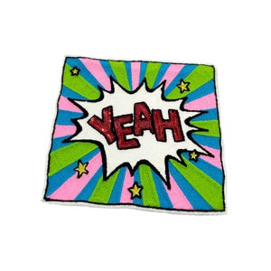 Puede incluir: Una almohada cuadrada colorida con un globo de diálogo blanco que dice "Yeah!" en lentejuelas rojas. El fondo es un patrón a rayas rosa, verde y azul con estrellas doradas.