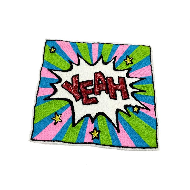 Puede incluir: Una almohada cuadrada colorida con un globo de di&aacute;logo blanco que dice "Yeah!" en lentejuelas rojas. El fondo es un patr&oacute;n a rayas rosa, verde y azul con estrellas doradas.