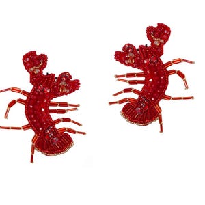 May include: Two red beaded lobster brooches. The brooches are intricately designed with small beads, creating a detailed representation of lobsters. The claws and legs are extended, and the overall design is decorative.