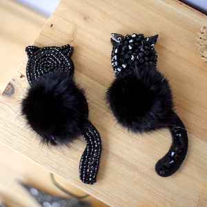 May include: Two black cat brooches. Each cat has a fluffy black fur body, with beaded heads and tails. One cat's head is made of small black beads, the other's of larger, faceted black beads. The brooches are on a wooden surface.