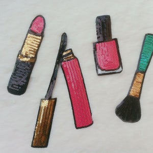 Può includere: Quattro articoli di trucco colorati fatti di paillettes, tra cui un rossetto rosa, uno smalto per unghie rosa, un pennello da trucco nero e un eyeliner dorato.
