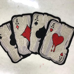 Pode incluir: Um emblema bordado com quatro cartas de jogar dispostas em leque. As cartas são o ás de paus, ás de ouros, ás de espadas e ás de copas. Cada carta tem um fundo cinza claro com uma borda preta espessa e bem definida ao redor.