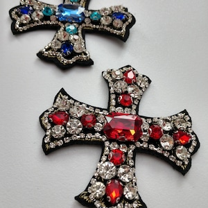 May include: Two black fabric cross-shaped patches with jeweled embellishments. One cross has red and clear jewels, the other has blue and clear jewels.