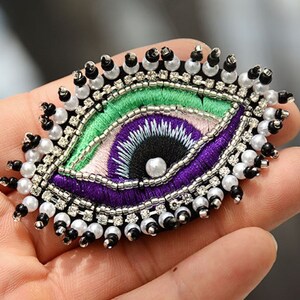 Könnte beinhalten: Ein gesticktes Auge in den Farben Lila, Grün und Weiß, mit schwarzen Perlen und weißen Perlen am Rand. Das Auge ist ein beliebtes Symbol für Schutz und Glück.