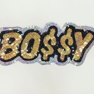 Op de afbeelding: Een pailletten patch met het woord "BOSS$Y" in gouden pailletten omlijnd met zwart, met een zilveren rand. De patch is ontworpen om op kleding of accessoires te naaien of te strijken.