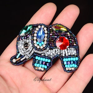 May include: A beaded elephant brooch with a black base, featuring colorful gemstones and beads. The elephant has a blue, green, and red gemstone design, with turquoise and silver bead accents. The word "Elephant" is written in cursive.