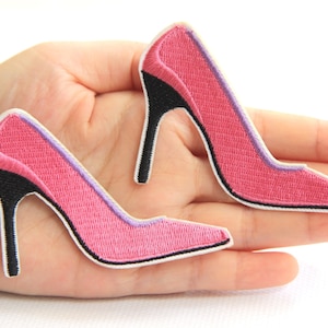 Peut inclure: Deux écussons brodés en forme d'escarpins roses avec des talons noirs et une bordure violette. Les écussons sont bordés de blanc. Ils sont conçus pour être cousus sur du tissu.
