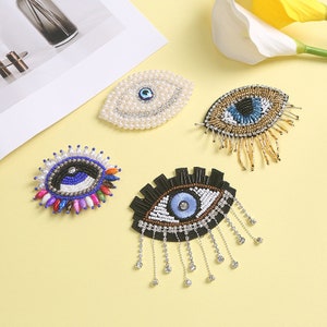 May include: Four different evil eye charms with beaded designs. The charms are in the shape of an eye with a blue centre and are surrounded by beads of various colours. The charms are on a yellow background.