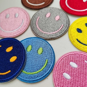 Könnte beinhalten: Eine Sammlung gestickter Smiley-Gesichter-Patches in verschiedenen Farben, darunter Rosa, Blau, Gelb, Rot, Braun und Grau. Jeder Patch zeigt ein lächelndes Gesicht mit kontrastierendem Faden für Augen und Mund. Diese Patches sind rund und haben eine strukturierte Optik.