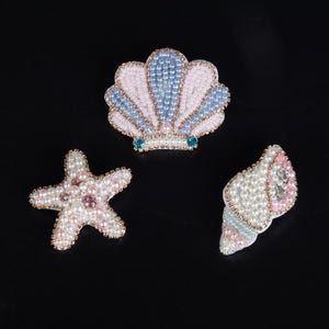 May include: Three beaded hair clips in the shape of a seashell, a starfish, and a seashell. The seashell hair clips are pink and blue, the starfish is pink, and the seashell is white and pink.