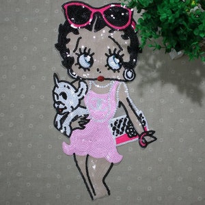 Può includere: Toppa in paillettes da stirare con un personaggio dei cartoni animati che indossa occhiali da sole, un vestito rosa e tiene un piccolo cane bianco. Il personaggio tiene una borsa a mano bianca e nera.