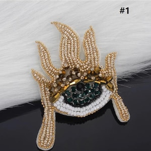 May include: A gold and white beaded eye with green rhinestones, surrounded by a beaded flame design. #1