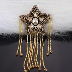 Könnte beinhalten: Ein goldener, sternförmiger Perlenbesatz mit einer Fransen aus goldenen Perlen. Der Stern ist mit klaren und braunen Strasssteinen verziert.