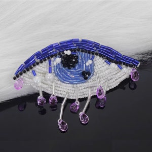 May include: A blue and white beaded eye with purple teardrop beads hanging from the bottom. The eye is surrounded by white beads and has a black pupil.